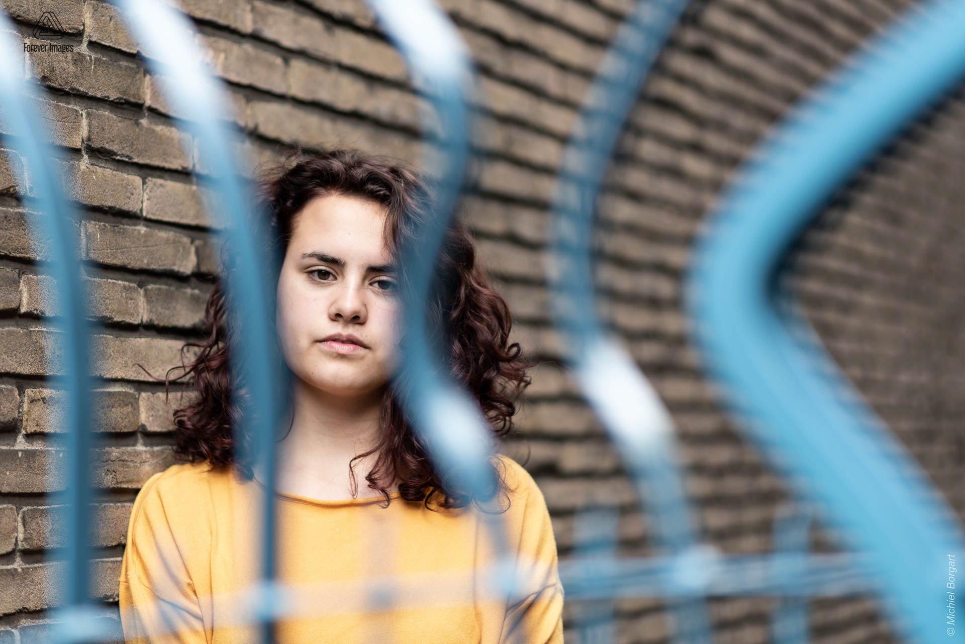 Portrait photo young lady against wall behind blue fence looking in camera | Tessa Holscher | Portrait Photographer Michiel Borgart - Forever Images.