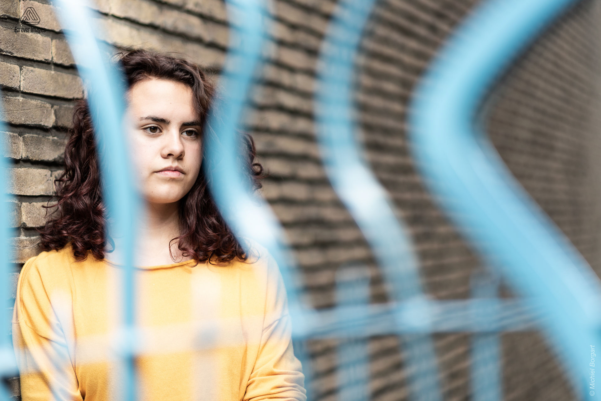 Portretfoto jonge dame tegen muur achter blauw hek kijkend opzij | Tessa Holscher | Portretfotograaf Michiel Borgart - Forever Images.