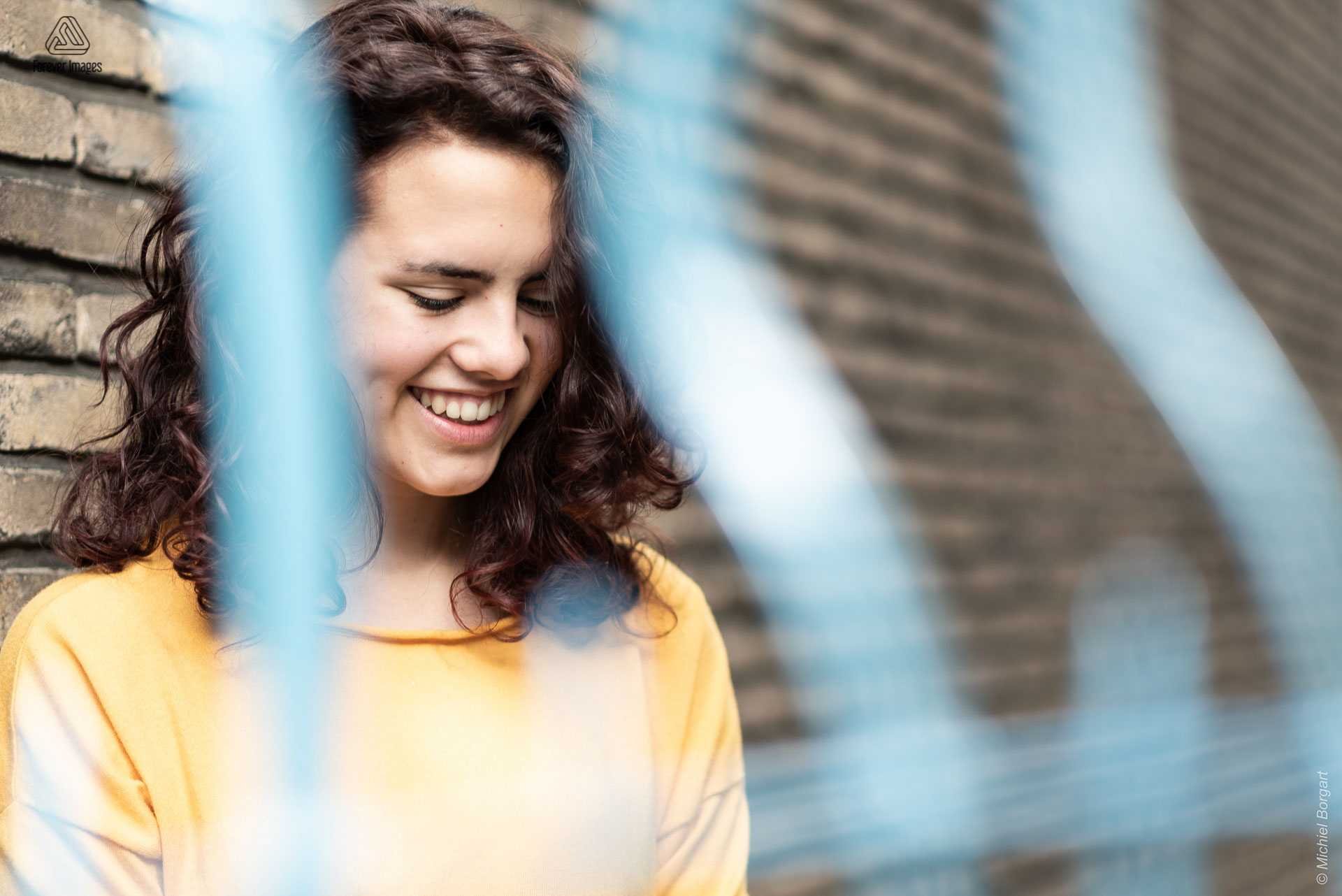 Portretfoto jonge dame tegen muur achter blauw hek lachend kijkend naar beneden | Tessa Holscher | Portretfotograaf Michiel Borgart - Forever Images.