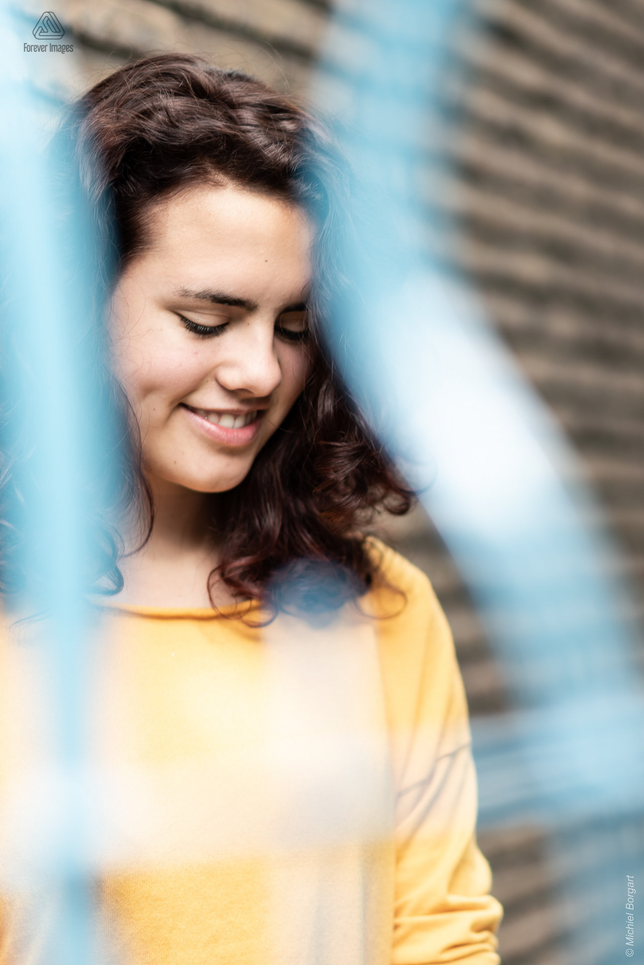 Portretfoto jonge dame tegen muur achter blauw hek zachte glimlach | Tessa | Portretfotograaf Michiel Borgart - Forever Images.