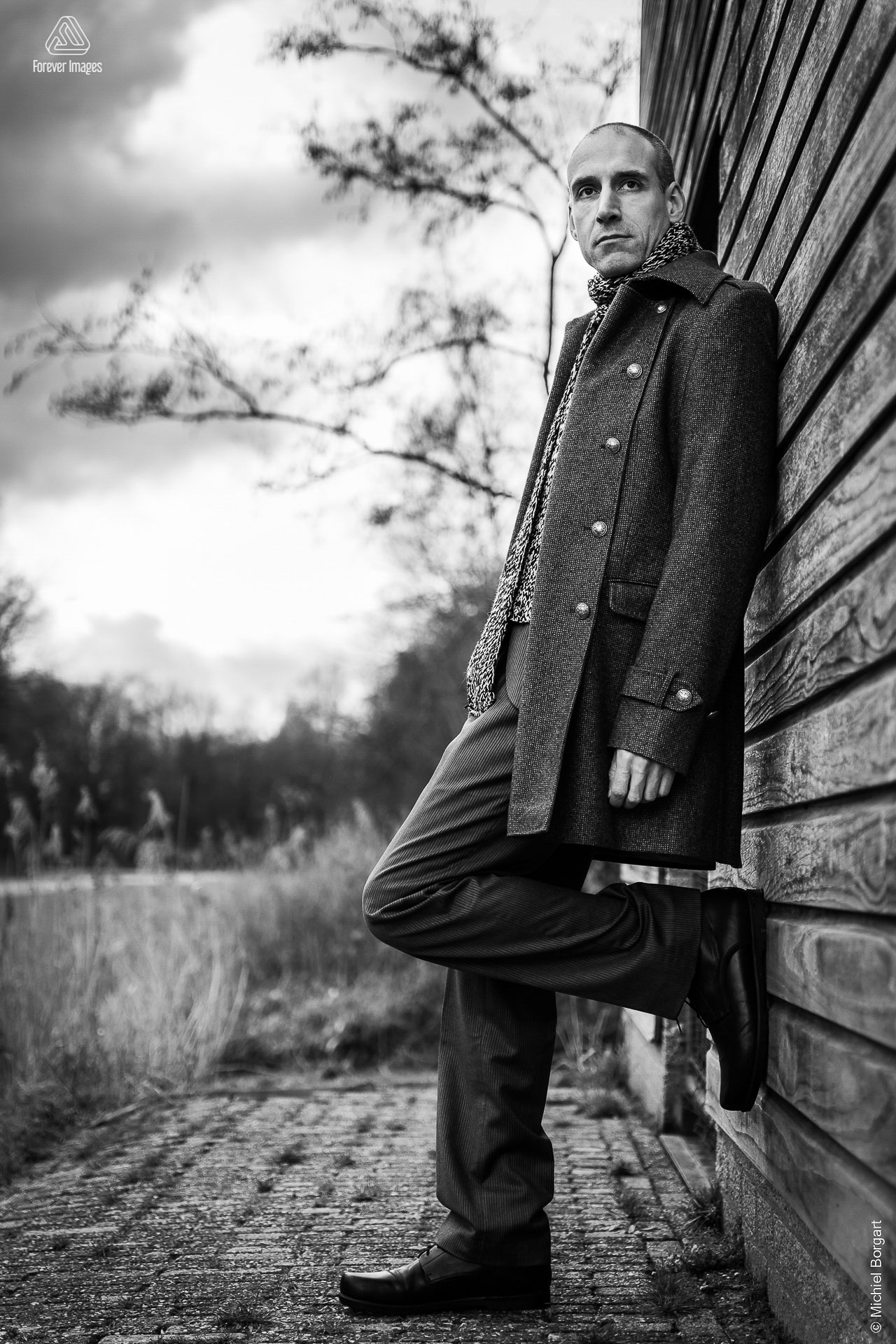 Portretfoto zwart-wit man met winterjas en sjaal tegen houten gebouw | Robin Het Twiske De Stootersplas | Portretfotograaf Michiel Borgart - Forever Images.